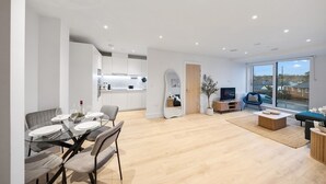 A modern living room with a glass dining table, grey upholstered chairs, a wooden coffee table, and a large window offering a view of the outside. - Comfy 2 BR+ Free Parking Gym, Game Rooms, & Cinema (London)