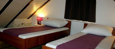 Een kleine, knusse kamer met twee houten bedden, een nachtkastje met een lamp en een raam met gordijnen.