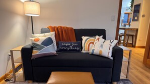 Un salón con un sofá negro, un reposapiés marrón y varios cojines de colores.