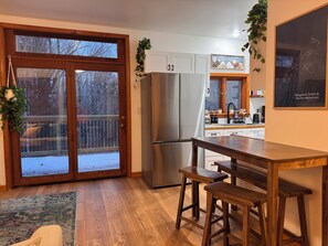 A kitchen with a large refrigerator, wooden stools, and a view of a snowy deck through glass doors. - Little House at Burke Basecamp--Ride-In Ride-Out Kingdom Trails + AC (Burke)