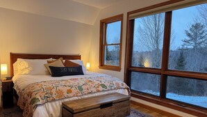 Un dormitorio con una cama grande, una mesita de noche, un cofre de madera y una ventana con vistas a árboles.