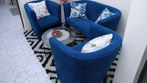 Un ensemble de canapés bleu agrémenté de coussins à motifs floraux, une table basse en verre avec des bonbons colorés, et un tapis à motifs posé sur un sol carrelé.