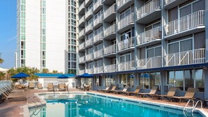 Un espace piscine doté de chaises longues et de parasols, entouré d’un immeuble à plusieurs étages avec des balcons.