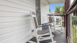 Une véranda avec deux fauteuils à bascule et une petite table, une terrasse en bois et un mur extérieur peint en blanc.