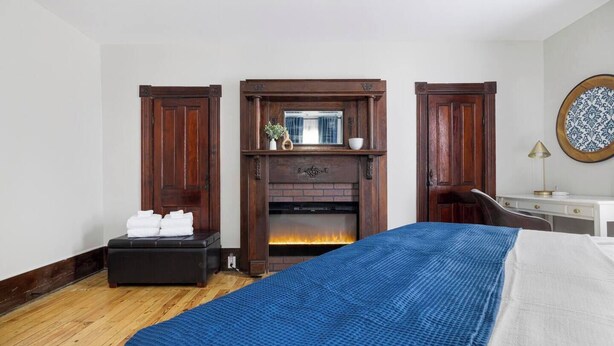 Une chambre avec une cheminée, un lit recouvert d’une couverture bleue, un banc en bois avec des serviettes et un bureau avec une lampe.