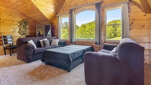 Ein gemütliches Blockhaus-Interieur mit Holzbalkendecke, großen Fenstern, einer Couch, einem Sessel und einem kleinen Tisch.