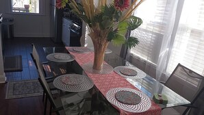 Een eetgedeelte met een glazen tafel, een rode tafelkleed en een vaas met bloemen. Achtergrond is een keuken met een koelkast en een raam.
