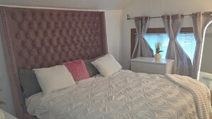 Een slaapkamer met een gestoffeerd hoofdeinde, een groot bed met witte beddengoed, een nachtkastje met een lamp en een raam met dunne gordijnen.