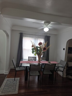 Un coin repas avec une table en verre, des chaises, un vase de fleurs et un ventilateur de plafond.