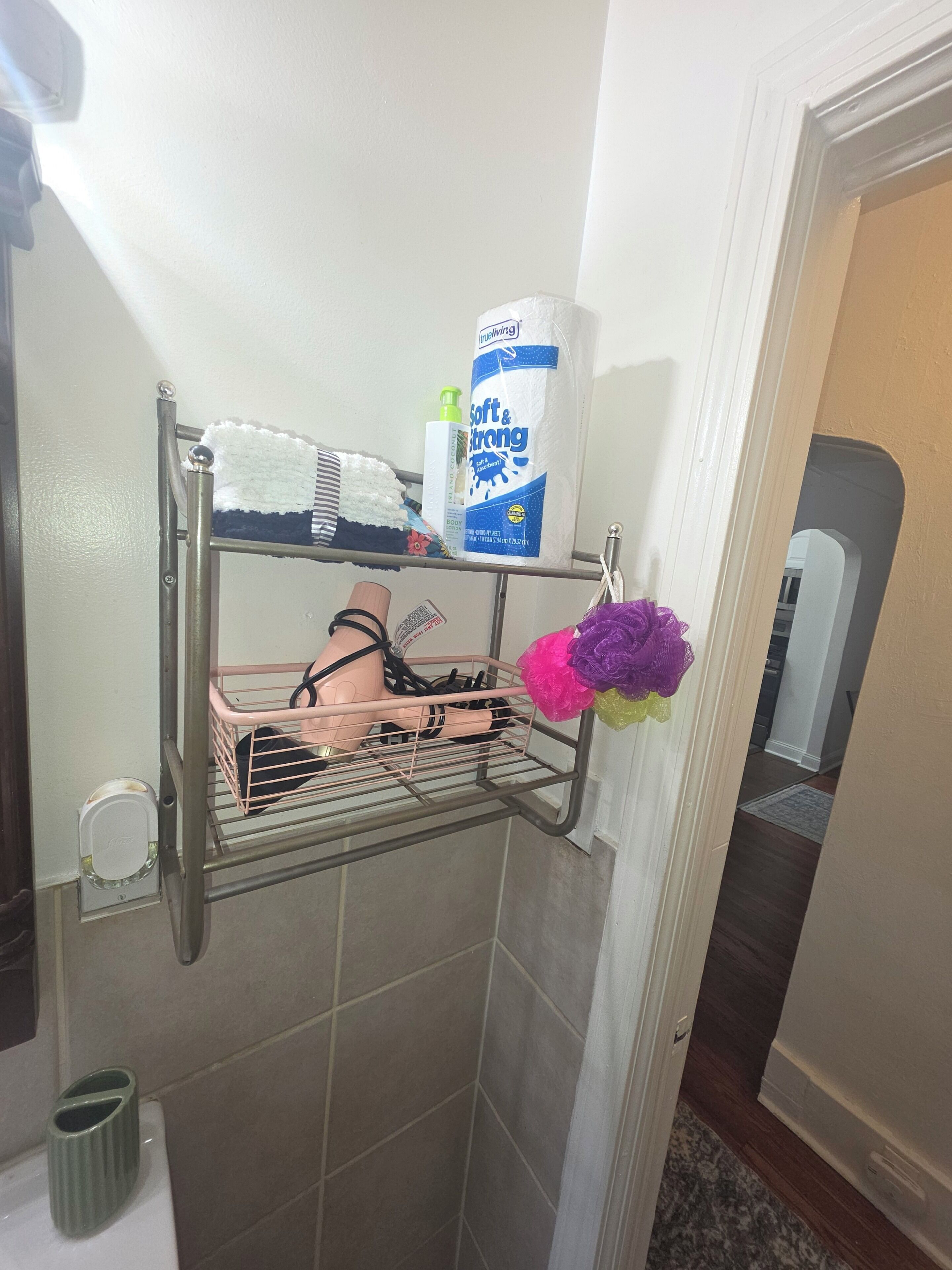 Une salle de bain avec un porte-serviettes sur lequel est posée une serviette rayée, une bouteille de gel douche doux et efficace, et un panier rose contenant un objet noir.