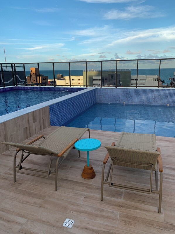Un área de piscina en la azotea con dos sillones de descanso y una mesita pequeña, rodeada por una baranda de cristal con vistas a la ciudad y al mar.