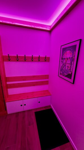 Une pièce avec un mur rose, un banc muni de crochets, un tableau encadré et un tapis noir sur le sol.