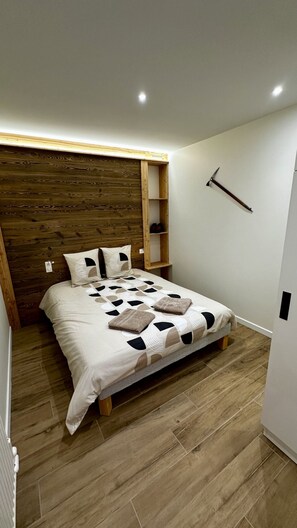 Une chambre à coucher avec une tête de lit en bois, un lit recouvert d’une couvre-lit à motifs géométriques, une table de chevet en bois et une étagère fixée au mur.