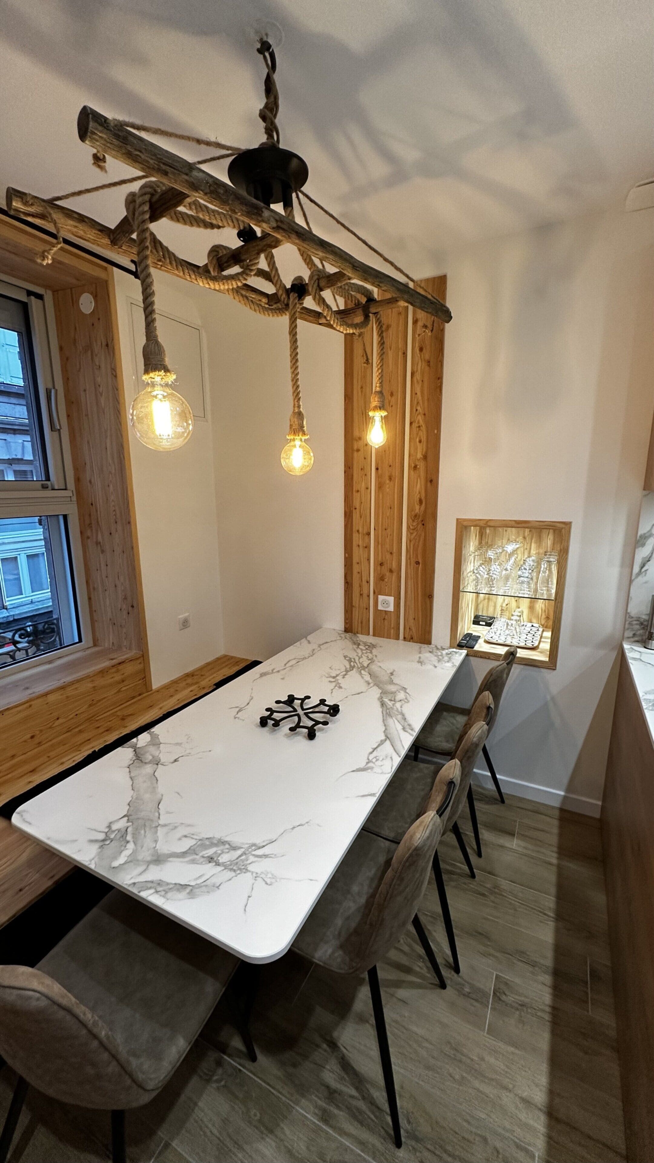 A dining area with a marble table, wooden chairs, a rope chandelier, and a view of the city through a window.