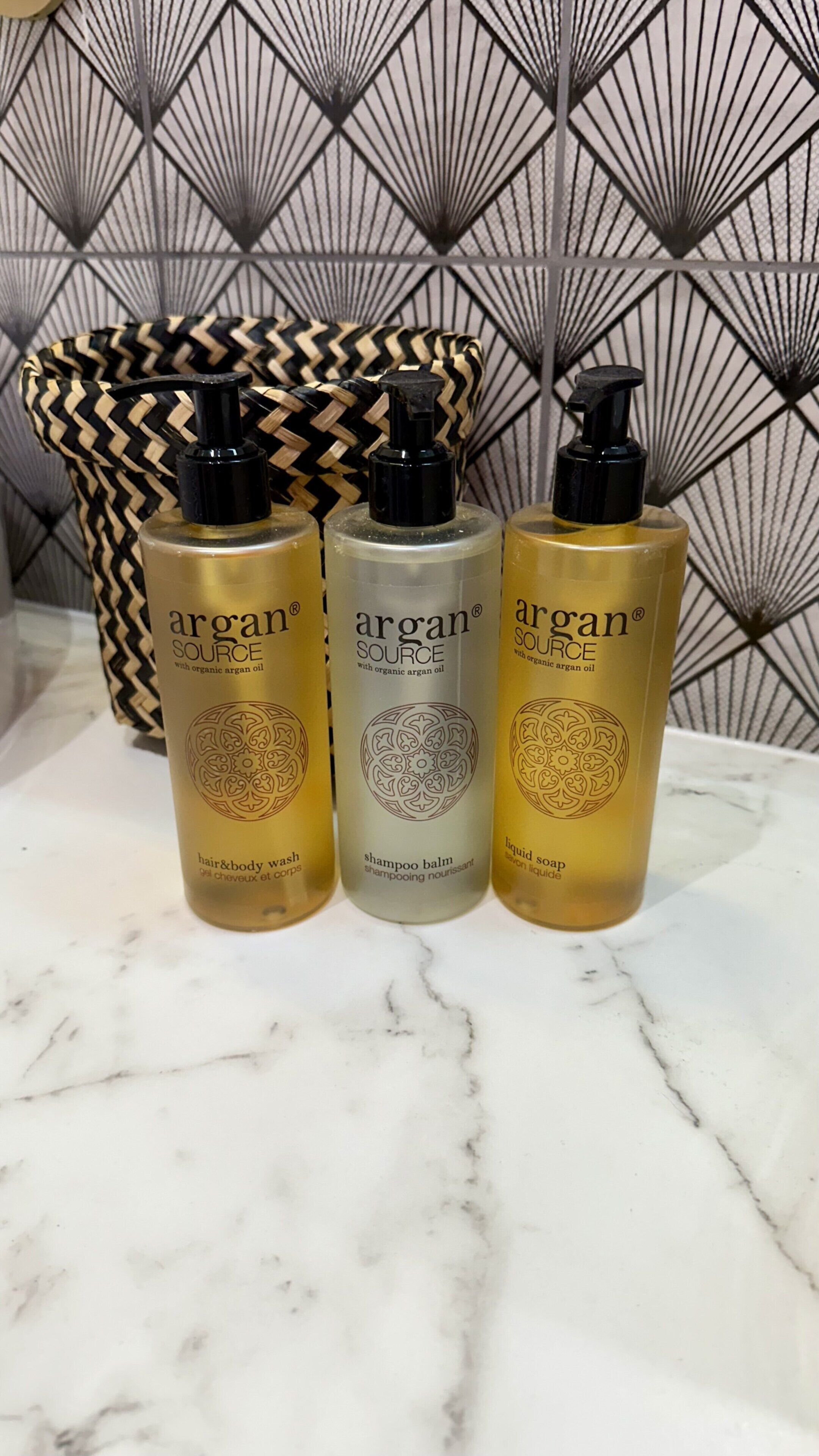 Drei Flaschen Argan Source auf einer Marmoroberfläche mit gemustertem Hintergrund.