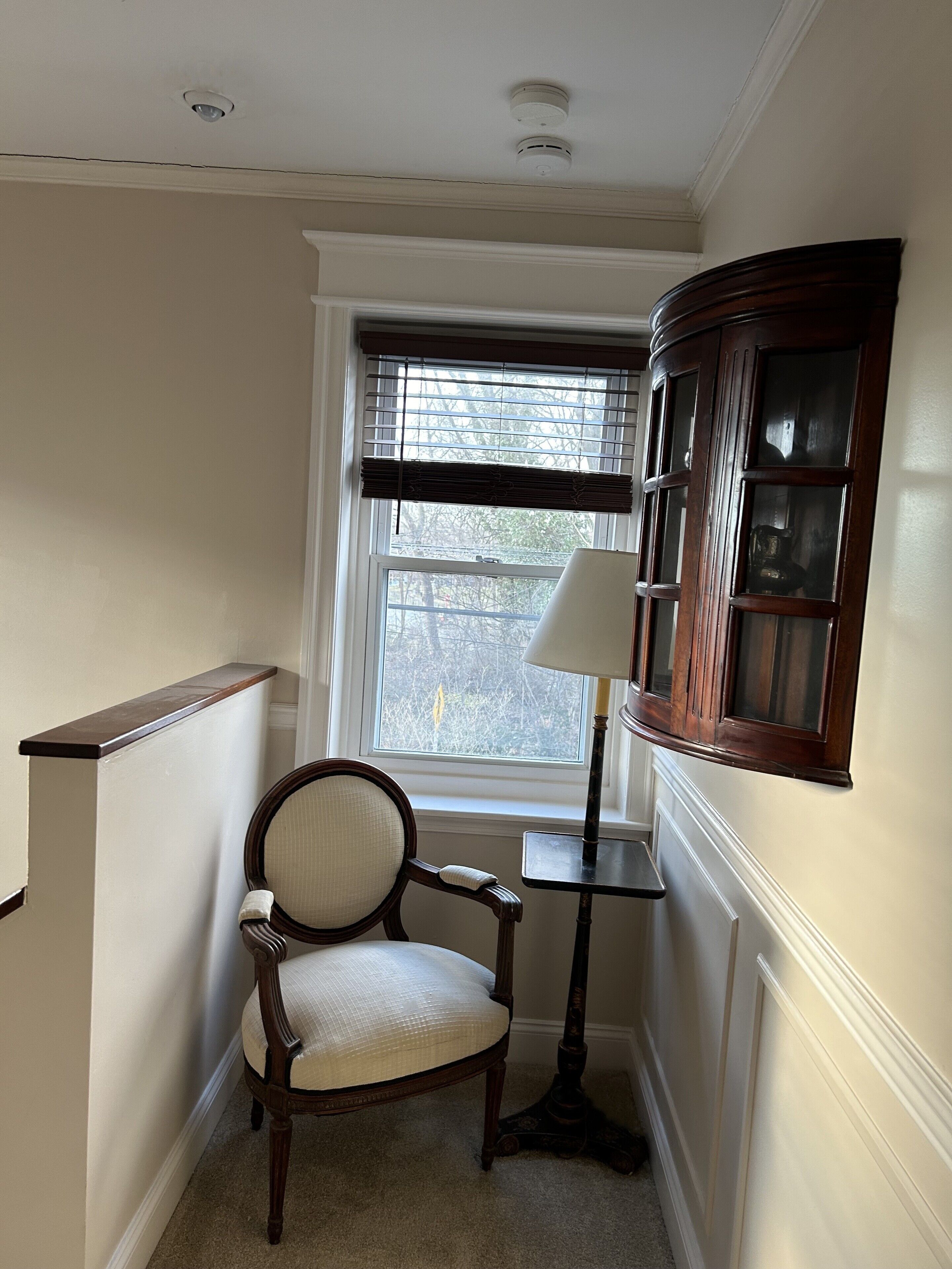 A corner room with a chair, a lamp, a window with blinds, and a wooden cabinet.