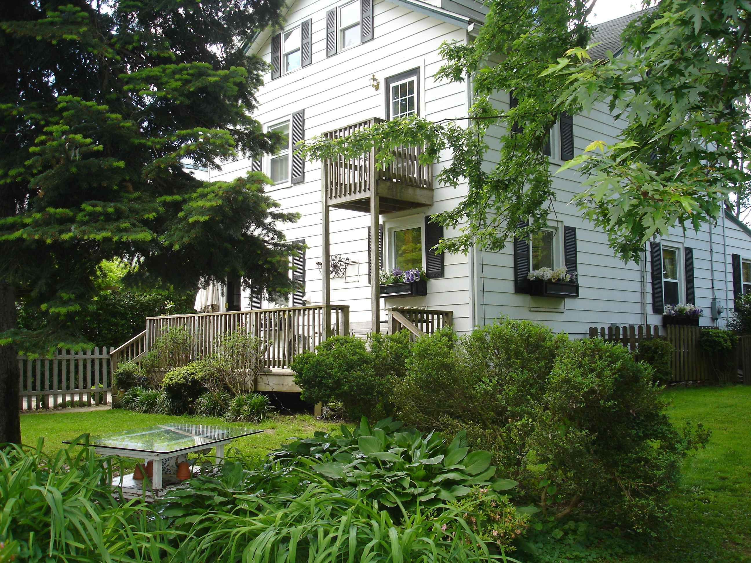 A white two-story house with a wooden deck, a small garden with various plants, and a glass table.