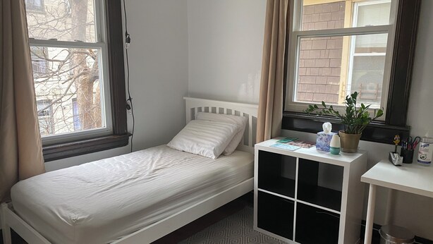 Habitación individual con una cama blanca, una mesita de noche con una planta y una ventana con cortinas.