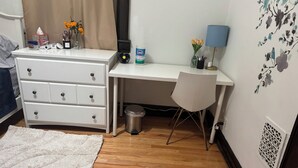 Un dormitorio con un aparador blanco, un escritorio con silla, un cubo de basura, una lámpara y una pared decorada con motivos florales.