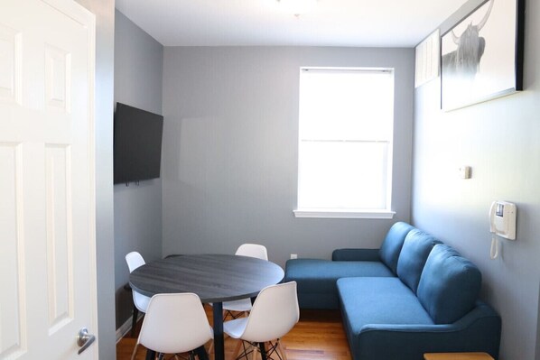 A room with a dark round table, white chairs, a blue sofa, a TV mounted on the wall, and a window with blinds. - Modern 1BR/1BA Apartment - Self Check-In (Belleville)