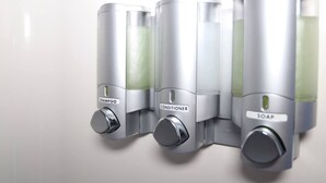 Ein wandmontierter Spender mit drei Fächern, beschriftet mit Shampoo, Conditioner und Seife.