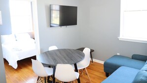Ein modernes Wohnzimmer mit rundem Tisch, weißen Stühlen, einem Flachbildfernseher, einem blauen Sofa und einem Bett mit weißer Bettwäsche.