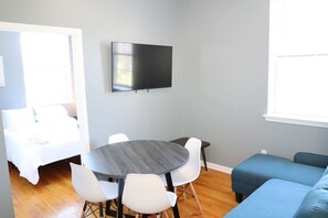 Ein modernes Wohnzimmer mit rundem Tisch, weißen Stühlen, einem Flachbildfernseher, einem blauen Sofa und einem Bett mit weißer Bettwäsche.
