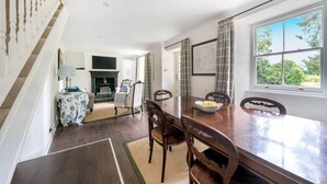A dining area with a wooden table and chairs, a fireplace, and a view of greenery outside. - Idyllic Waterside Georgian Cottage (Halberton)