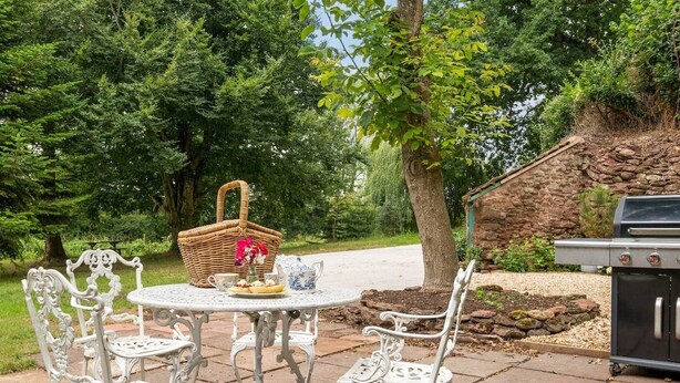 A patio with a round table and white chairs, a wicker basket with flowers, and a grill. - Idyllic Waterside Georgian Cottage (Halberton)