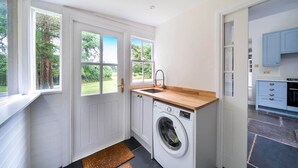 A compact kitchen with a washing machine, a sink, and a window overlooking a garden. - Idyllic Waterside Georgian Cottage (Halberton)