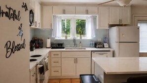 A kitchen with white cabinets, a window with curtains, a ceiling fan, and a refrigerator.