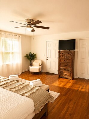 A bedroom with a bed, a ceiling fan, a television on a wooden dresser, and a chair.