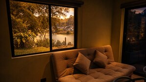 A room with a sofa facing a window overlooking a lake and trees. - Cabin with Jacuzzi in front of the Guatapé Lake. (Peñol)