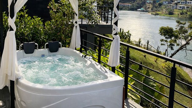 A hot tub with white curtains, a balcony with a view of a lake and mountains, and a wooden pergola structure. - Cabin with Jacuzzi in front of the Guatapé Lake. (Peñol)