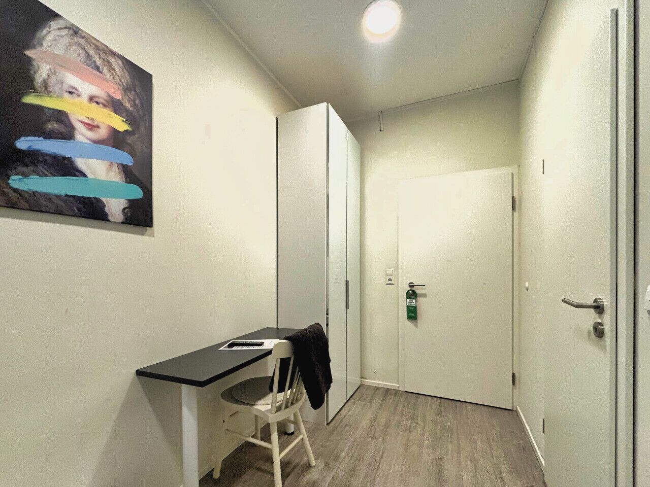 Una habitación pequeña y estrecha con escritorio y silla, perchero, puerta con extintor y un cuadro en la pared.