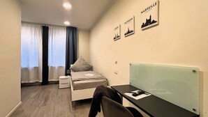 Une chambre d’hôtel de taille modeste, comprenant un lit, un bureau et une chaise. Deux tableaux encadrés ornent le mur, l’un représentant Lisbonne et l’autre, Bangkok.