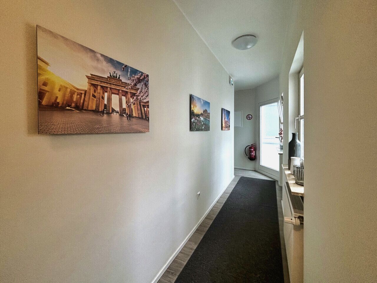 A hallway with a large painting of the Brandenburg Gate, a small window, and a door with a fire extinguisher.