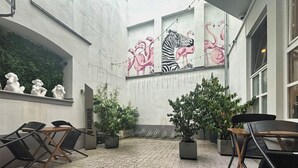 Un área de estar al aire libre con sillas y mesas, plantas en macetas y un mural con flamencos y una cebra en la pared.