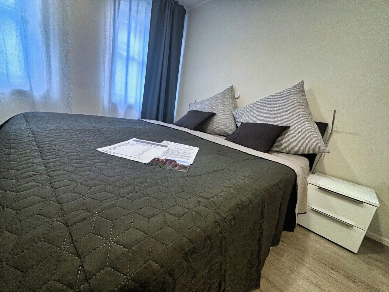 Una cama bien hecha con cubrecama acolchado, una mesita de noche con lámpara y una ventana con cortinas.