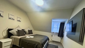 Un dormitorio con cama, mesita de noche, televisor y ventana con cortinas.