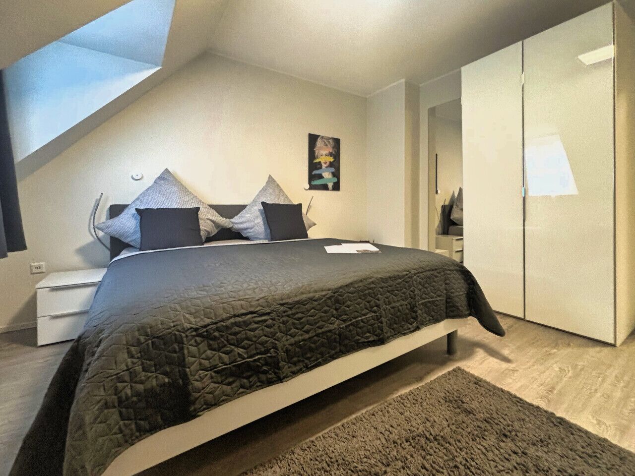 Un dormitorio con cama, mesita de noche, armario y tragaluz.