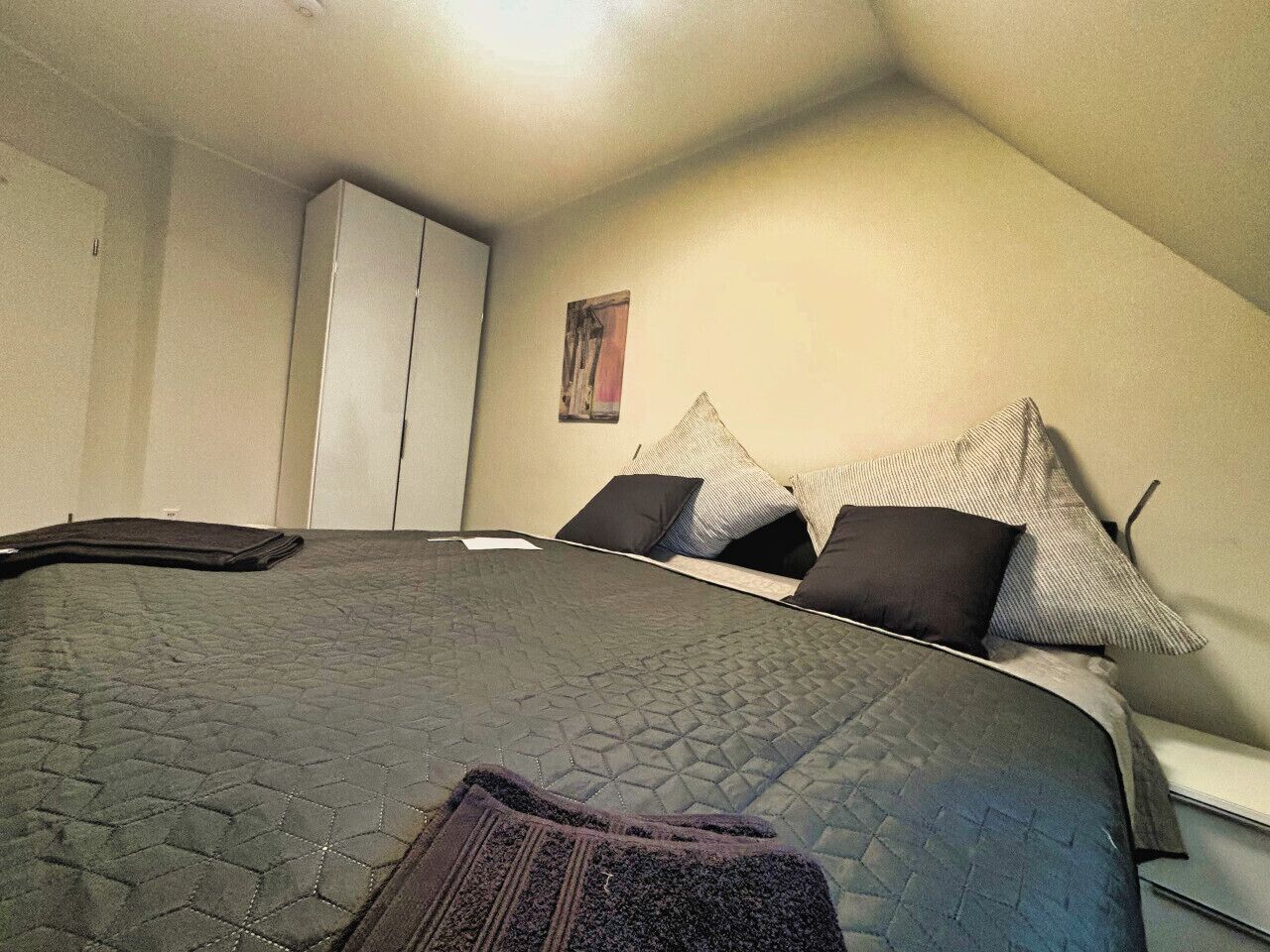 Un dormitorio con cama, almohadas, manta, armario y un cuadro en la pared.