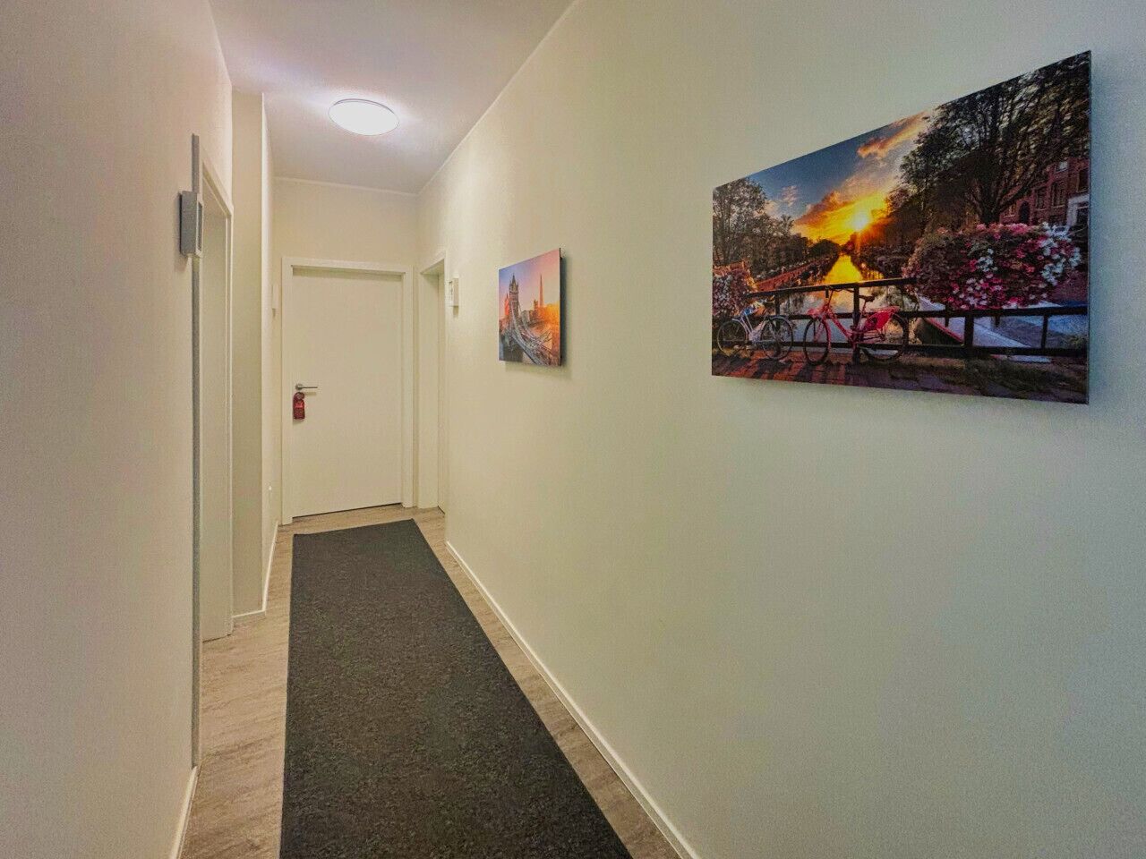 Un pasillo con alfombra oscura, paredes blancas y dos cuadros que representan paisajes.