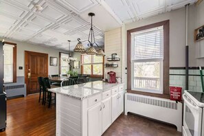 A kitchen with white cabinetry, a marble island, pendant lighting, and a large window with blinds. - Center House 1901 | Walk to Broadway | Parking (Jim Thorpe)