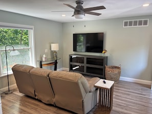 A modern living room with a reclining sofa, a flat-screen TV, a ceiling fan, and a window with a view of trees.