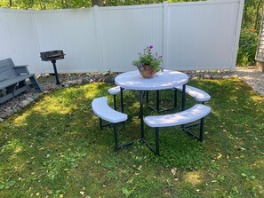 A round outdoor table with four white benches, a potted plant with purple flowers, and a white fence in the background.