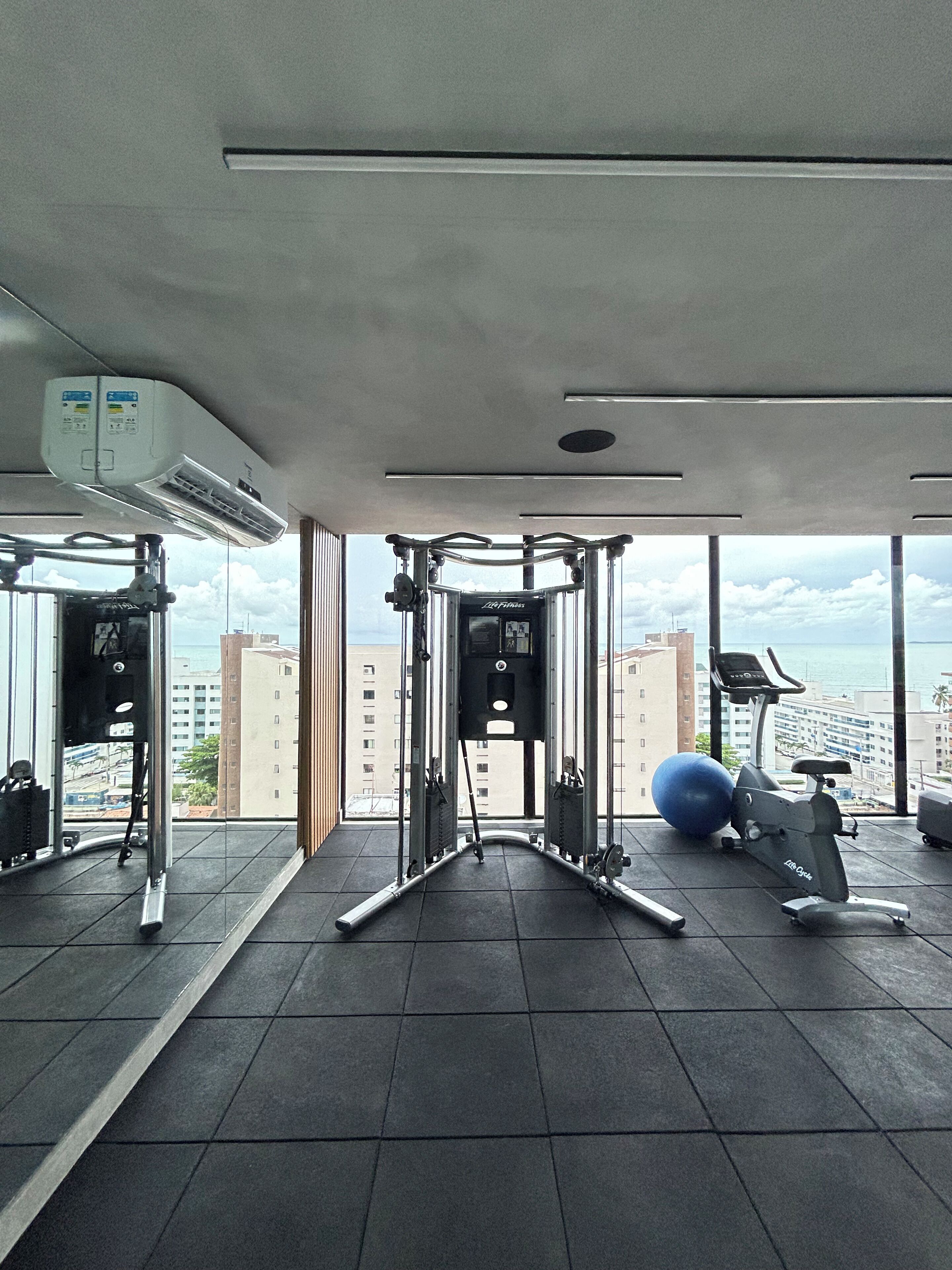 Un gimnasio con equipo de ejercicio, un ventanal y vistas a edificios.