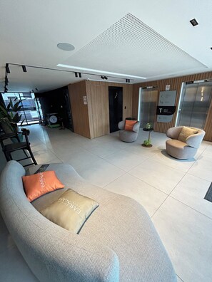 A modern living room with a sofa, armchairs, and a coffee table. There is a wooden panel wall, a door, and an elevator. - Melhores Flats - Surreal (Cabedelo)