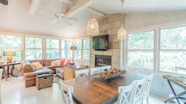A spacious living room with a wooden dining table, white chairs, a stone fireplace, and large windows offering a view of the outdoors. - Walk to white sand private beach of Oakwood Park - 4 bed cottage steps to lake. (Grand Bend)