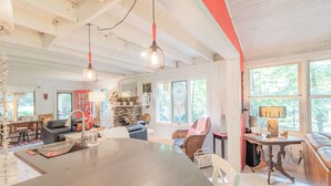 A spacious living room with a large central table, a fireplace, and a view of the outdoors through large windows. - Walk to white sand private beach of Oakwood Park - 4 bed cottage steps to lake. (Grand Bend)